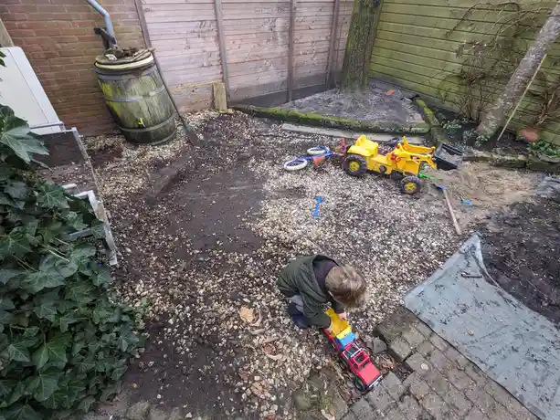 A young child in a green jacket is crouched in a backyard filled with gravel, dirt, and scattered construction toys…