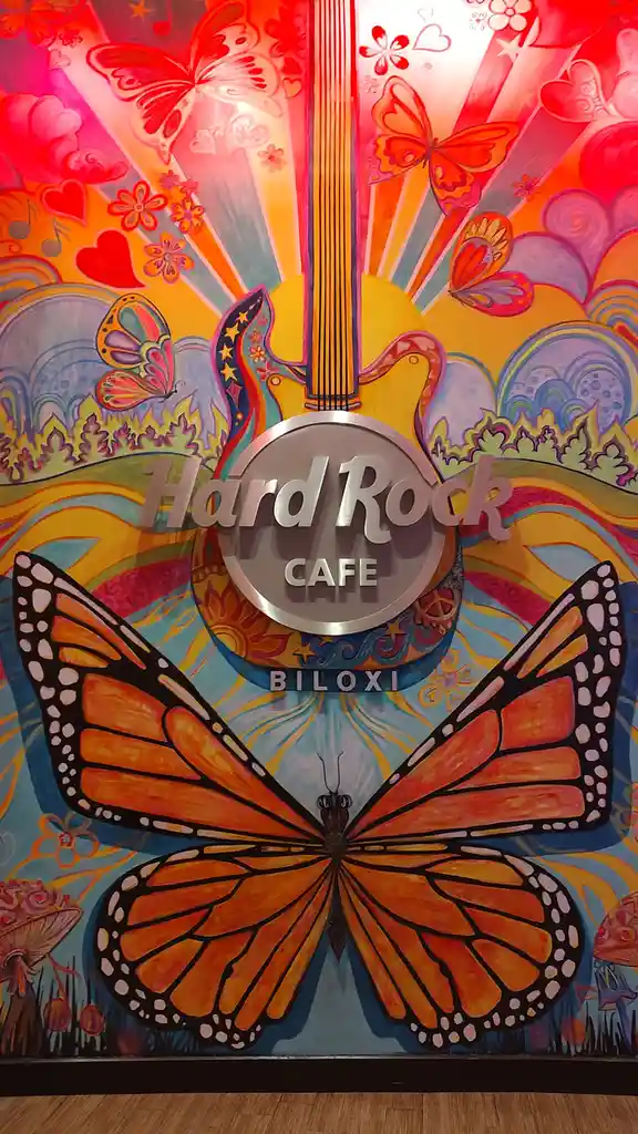 A vibrant, psychedelic mural features a large orange monarch butterfly at the bottom, with a yellow electric guitar rising…