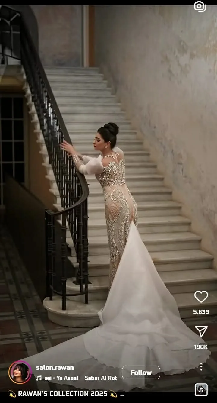 A woman in an ornate, beaded, floor-length white gown with a long train stands gracefully on a marble staircase, her hand…