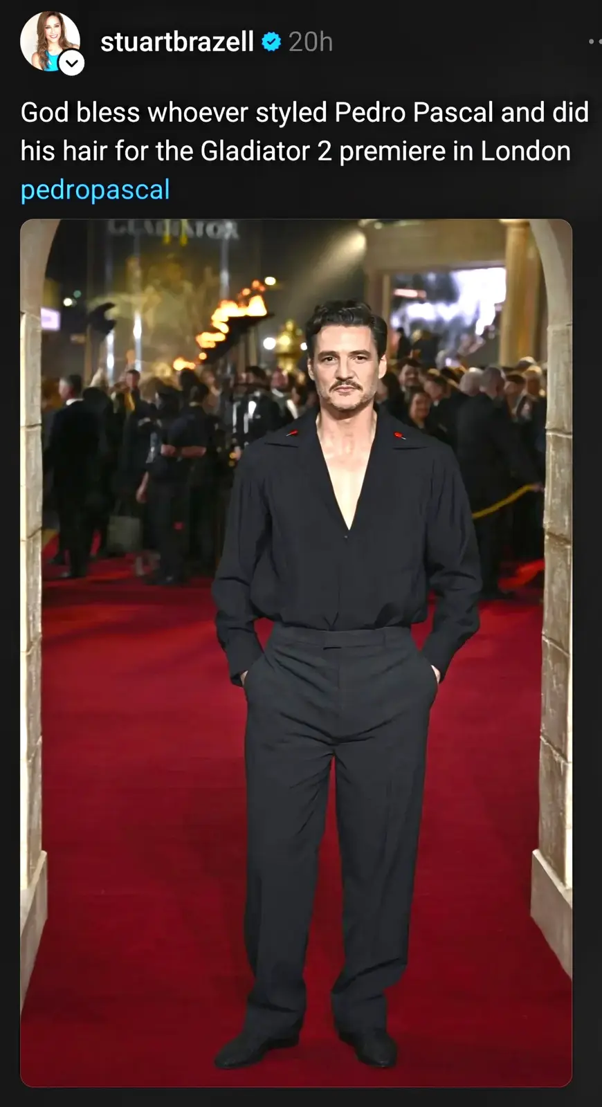 A man with dark hair and a mustache stands on a red carpet wearing a black, low-cut, button-up outfit with his hands in his…