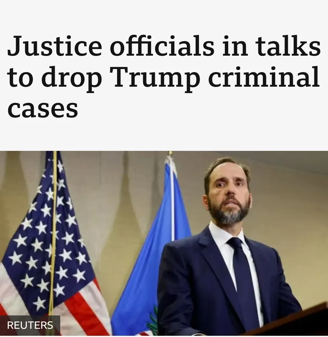 A man in a dark suit and tie stands at a podium with an American flag and another blue flag behind him. The overlaid text…