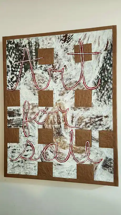 A handmade, rustic-looking artwork hangs on a wall, featuring the words 'Art for sale' outlined in red against a textured…