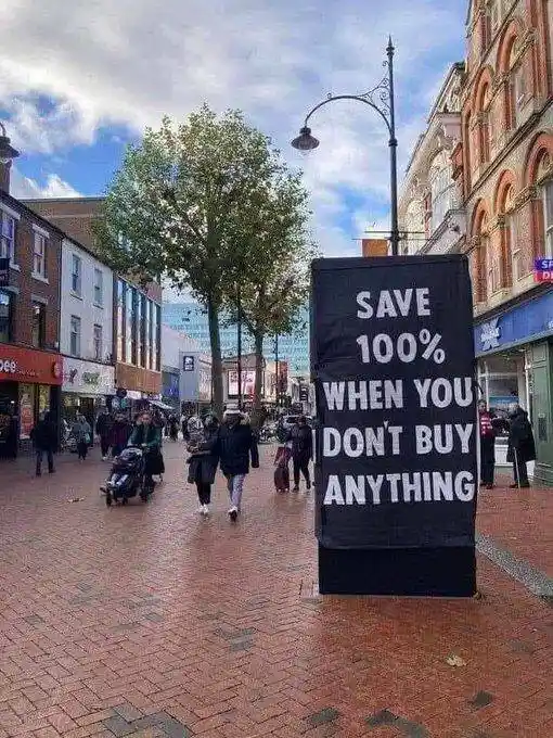 A black promotional banner stands on a brick pedestrian street, displaying white text that reads 'SAVE 100% WHEN YOU DON'T…