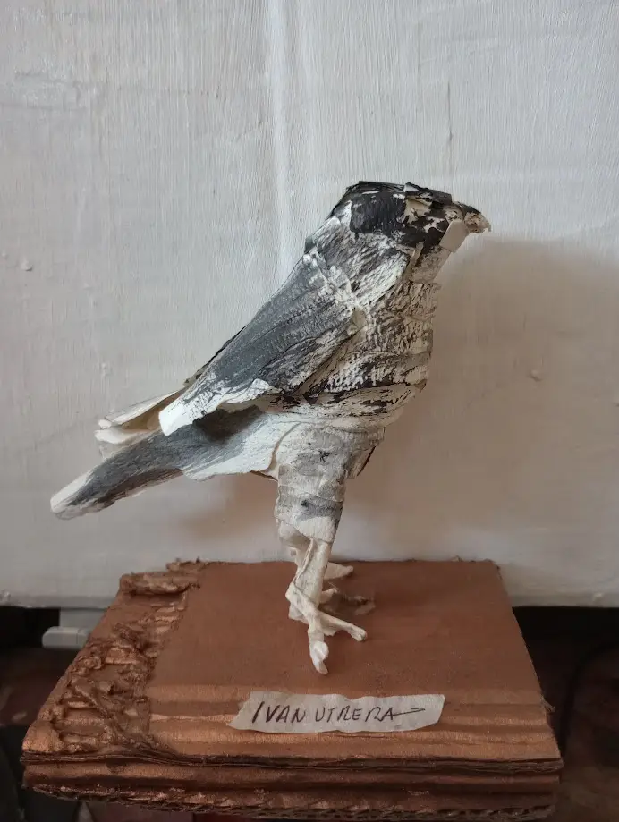 A handmade sculpture of a bird, possibly a crow or raven, crafted from layered birch bark or similar material, standing on a…