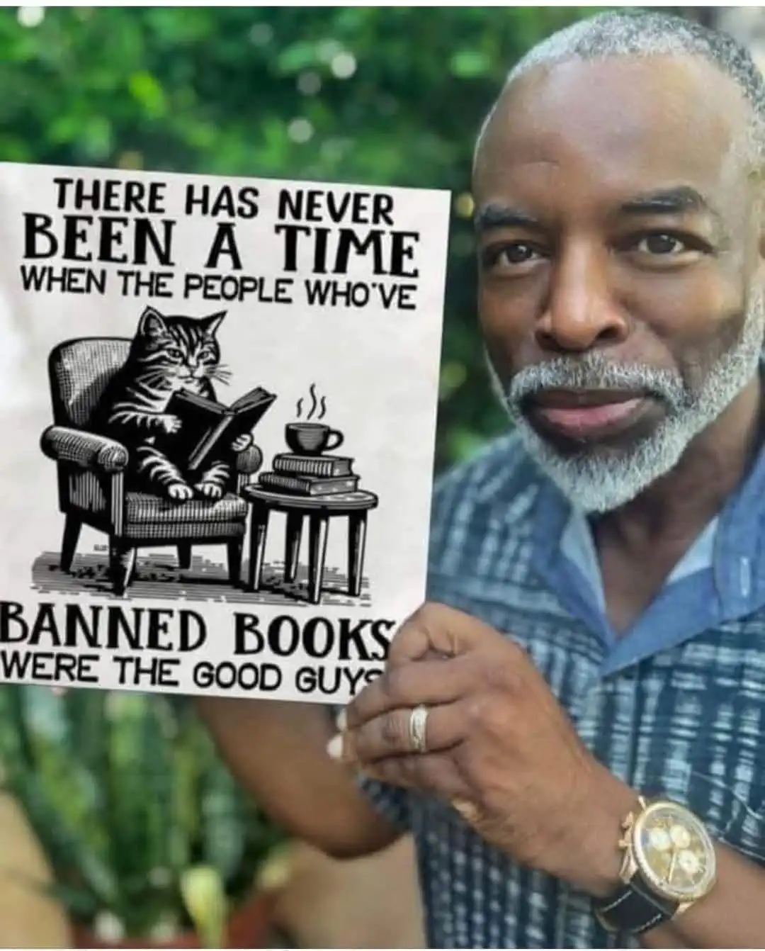 A man with gray hair and a beard holds up a sign that features a black-and-white illustration of a cat reading a book in an…