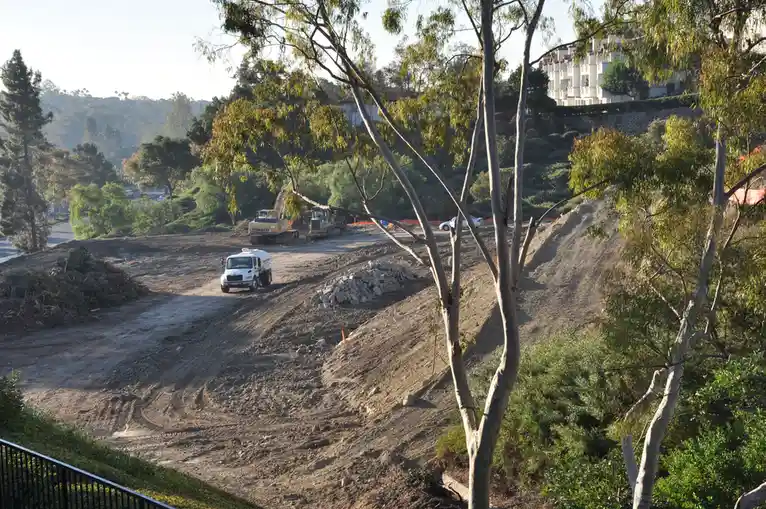 A photo of a dirt construction site with heavy machinery, including a Komatsu excavator and a white water truck, surrounded…