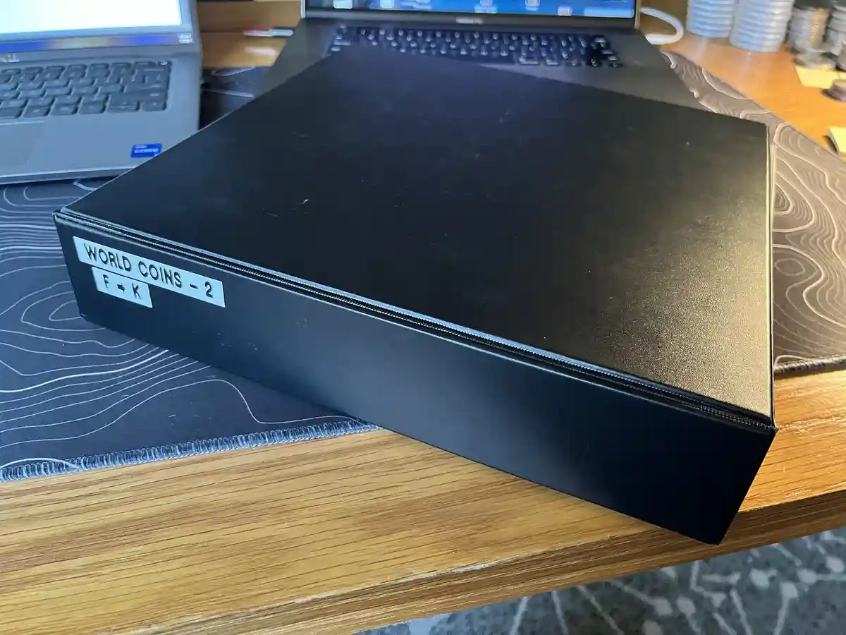 A black binder labeled 'WORLD COINS - 2' and 'F → K' sits on a wooden desk next to two laptops, humorously implying it…