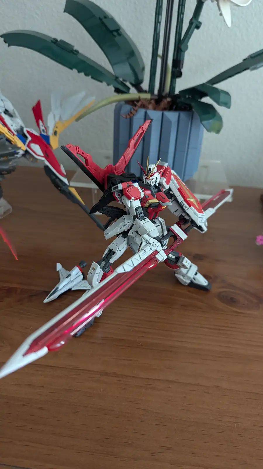 A red and white Gundam model stands on a wooden table holding a large red and white sword, with a potted plant featuring…