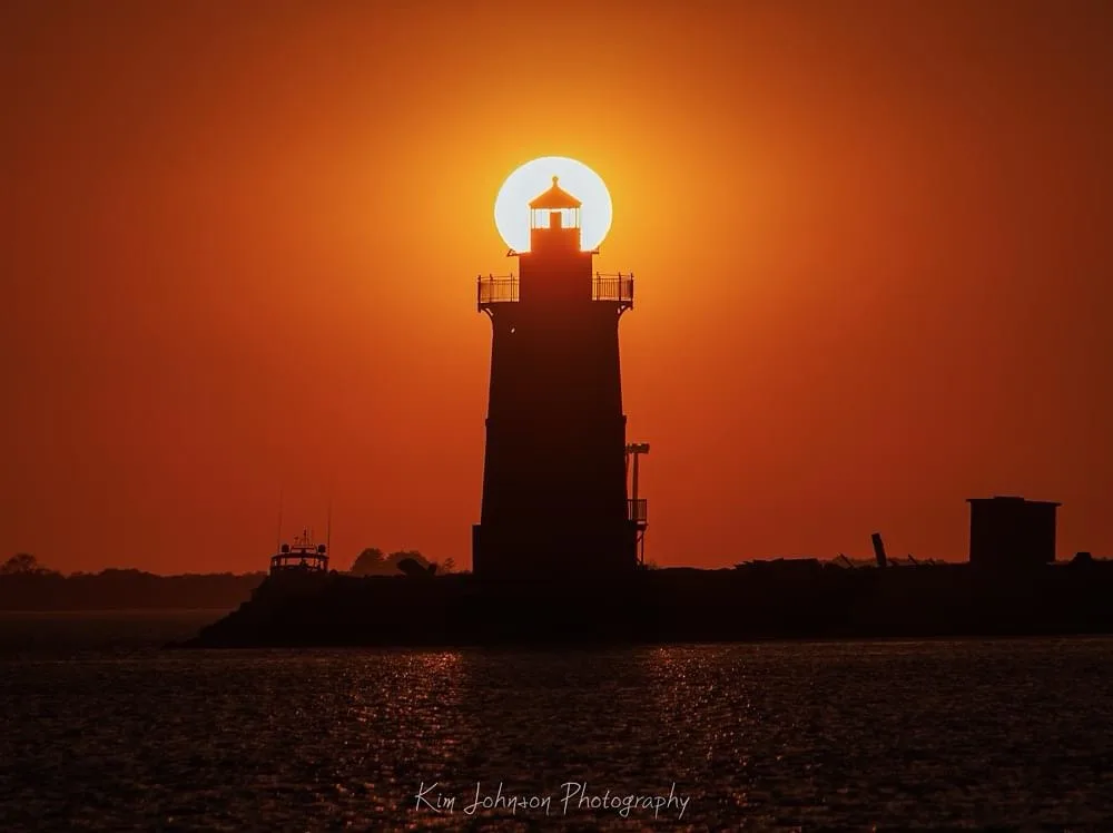 A lighthouse stands as a dark silhouette against a vibrant orange sunset, with the sun perfectly centered behind its lantern…