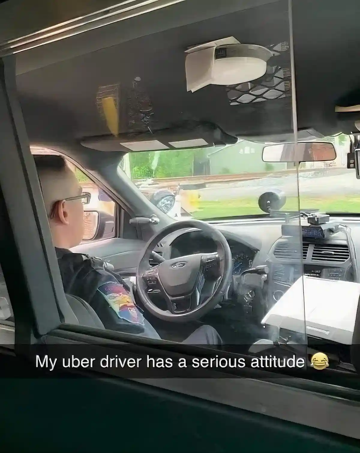 A passenger's view from inside a car shows a driver with a shaved head and glasses, looking straight ahead with a neutral…