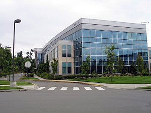 An exterior shot of a modern glass-and-concrete office building with a crosswalk in front, overlaid with the text 'this is…