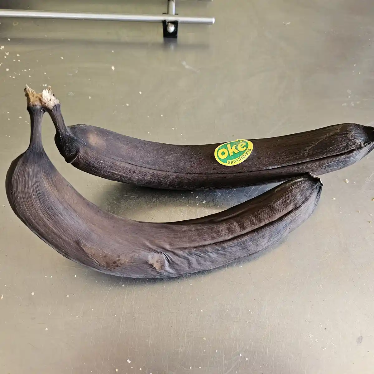 Two extremely overripe, dark brown bananas with shriveled tips sit on a stainless steel surface, one bearing a yellow and…