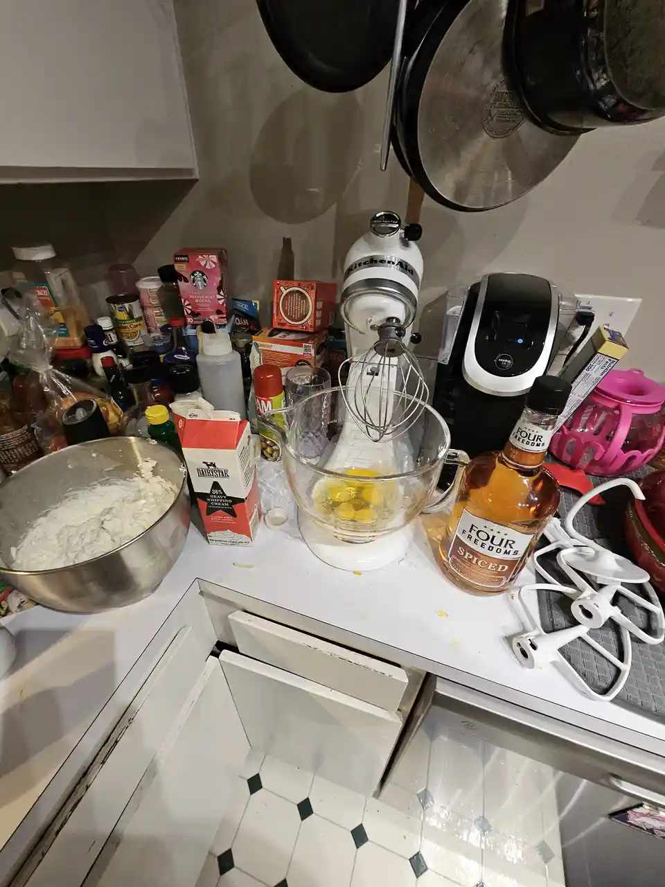 A cluttered kitchen counter features a white KitchenAid mixer with a glass bowl containing cracked eggs, a bottle of Four…