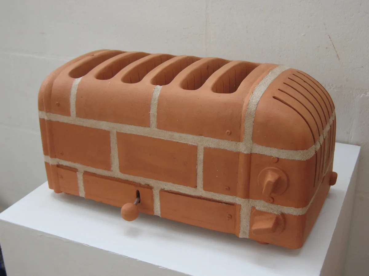 a toaster-shaped sculpture crafted from terracotta bricks and mortar, featuring slots, knobs, and a handle, displayed on a…