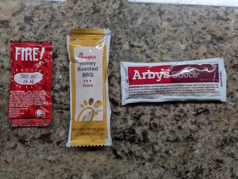 Three fast-food sauce packets lie side-by-side on a speckled countertop: a red Taco Bell 'FIRE!' sauce packet with 'THIS…