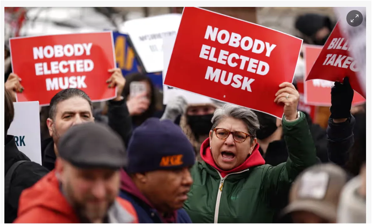 A crowd of protesters holds up red signs with bold white text reading 'NOBODY ELECTED MUSK'. A woman in a green jacket and…