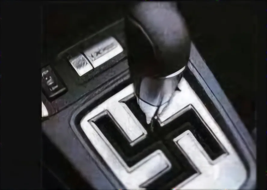 a close-up shot of a car's gear shift lever positioned over a gear pattern that has been altered to resemble a swastika…