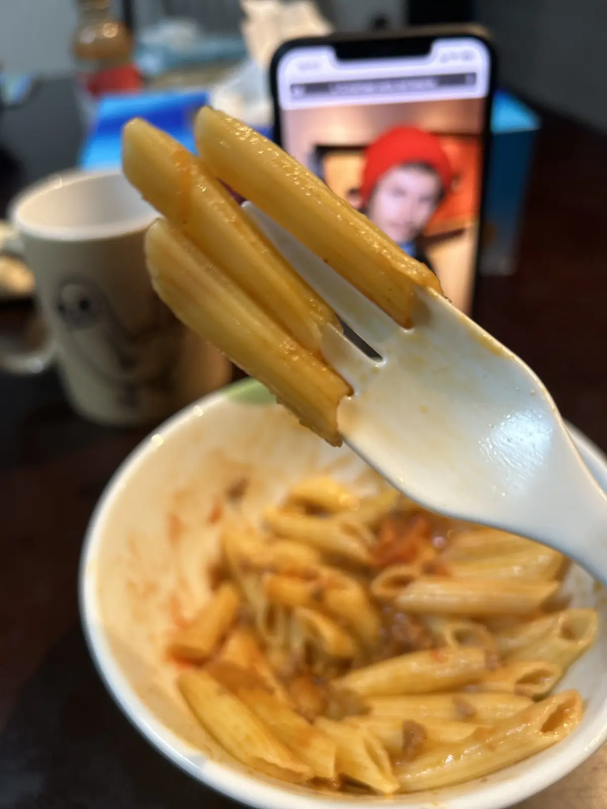 A close-up photo shows a white fork holding three penne pasta noodles coated in sauce, lifted from a nearly empty white…