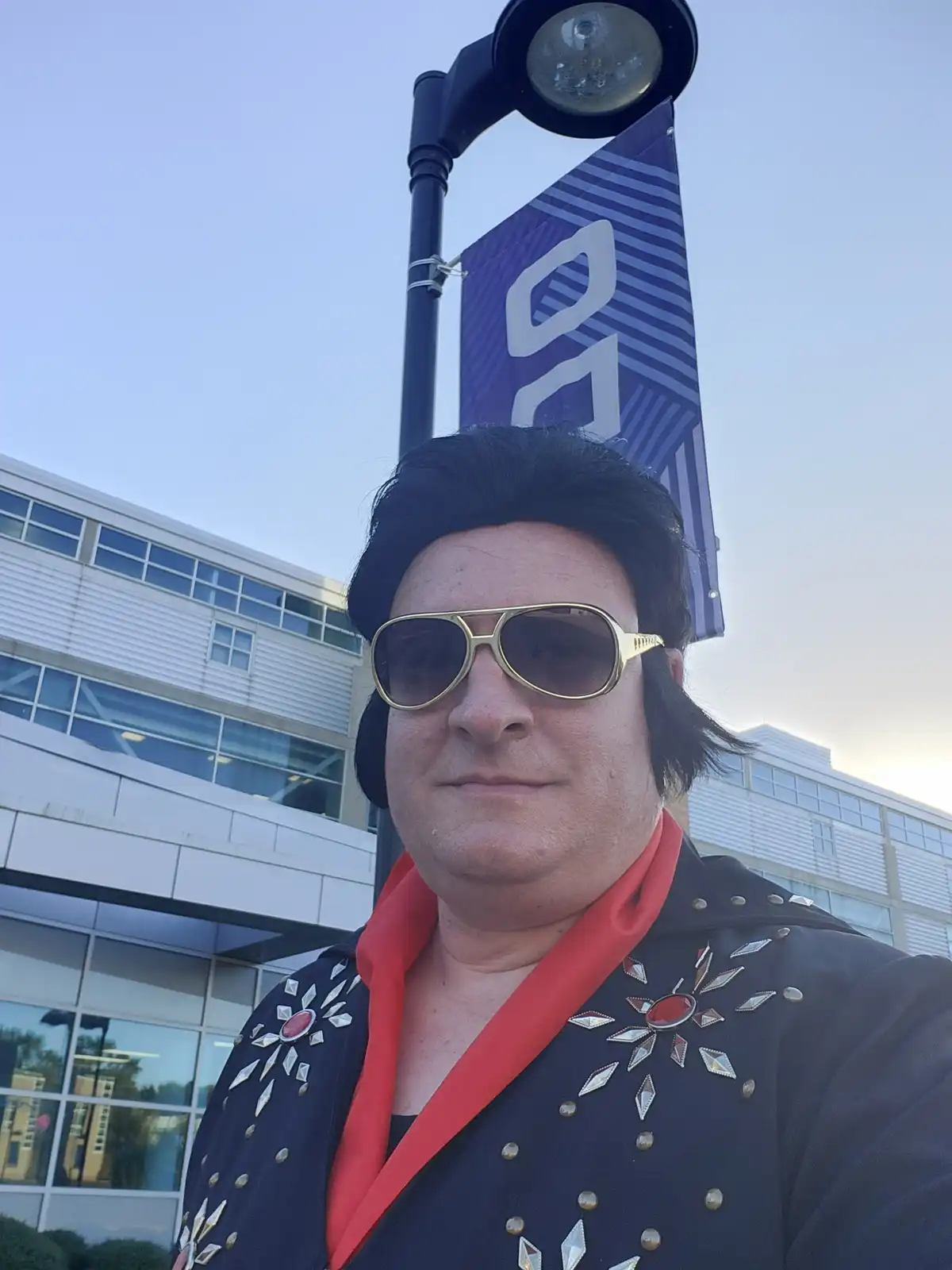 A person dressed as Elvis Presley, wearing a black sequined jacket with red lapels, gold-rimmed sunglasses, and a black wig…