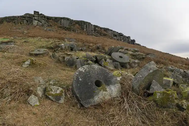 The image shows numerous large, round, flat stone millstones scattered across a grassy, rocky hillside under an overcast…