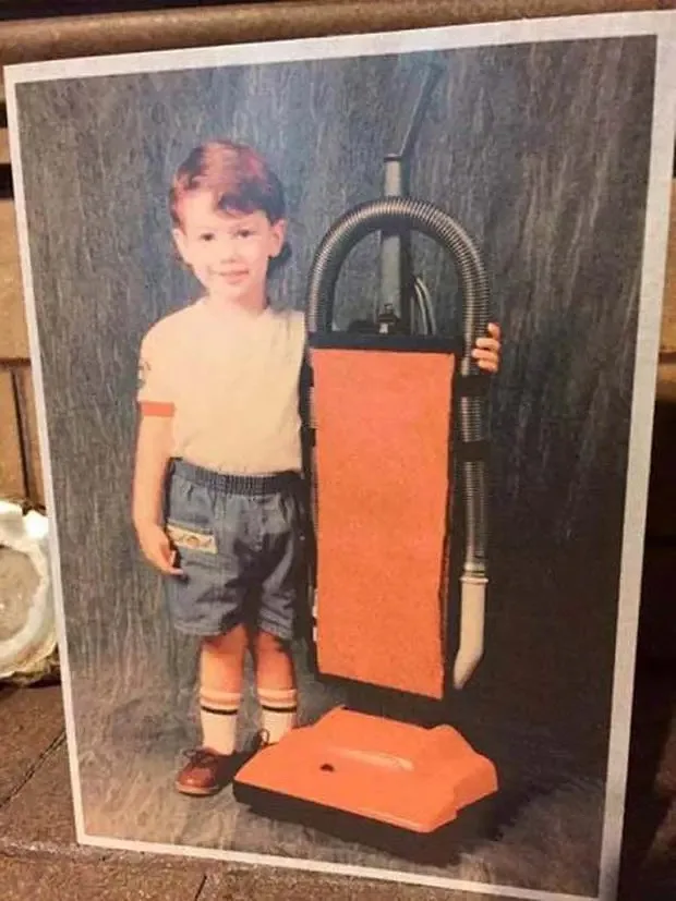 A vintage photograph shows a young child standing next to a large, orange upright vacuum cleaner, holding its hose. The…