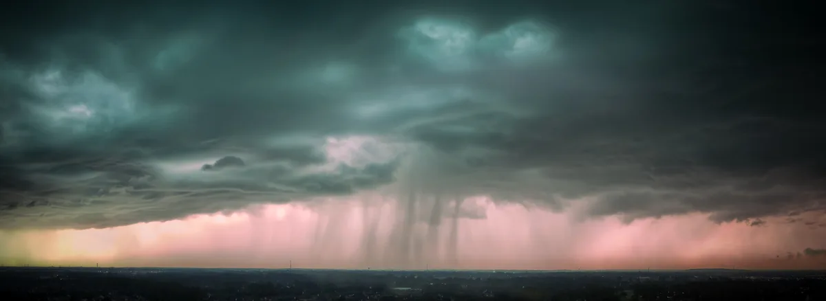 A panoramic photograph shows a dramatic, dark storm cloud formation looming over a distant cityscape at dusk. Rain is…