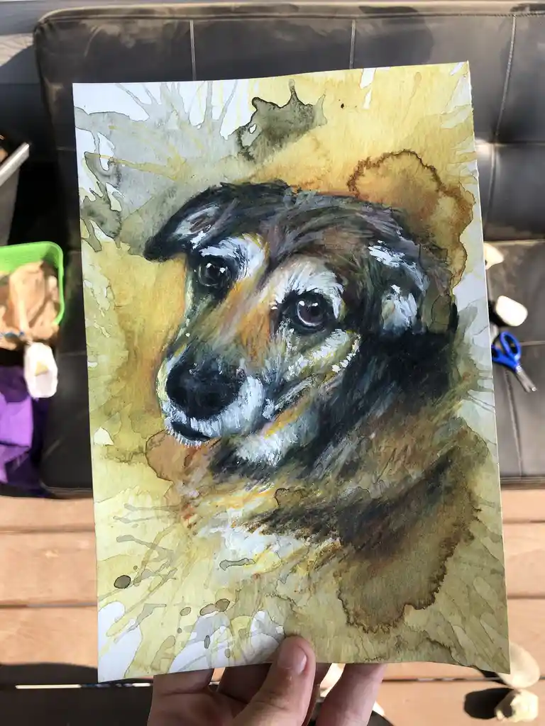 A hand holds up a watercolor painting of a dog's face, which has been rendered with dark, smudged markings around the eyes…