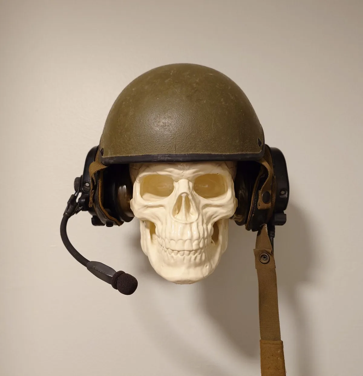 A white plastic skull wearing an olive-drab military helmet with attached black earphones and a microphone boom, mounted…