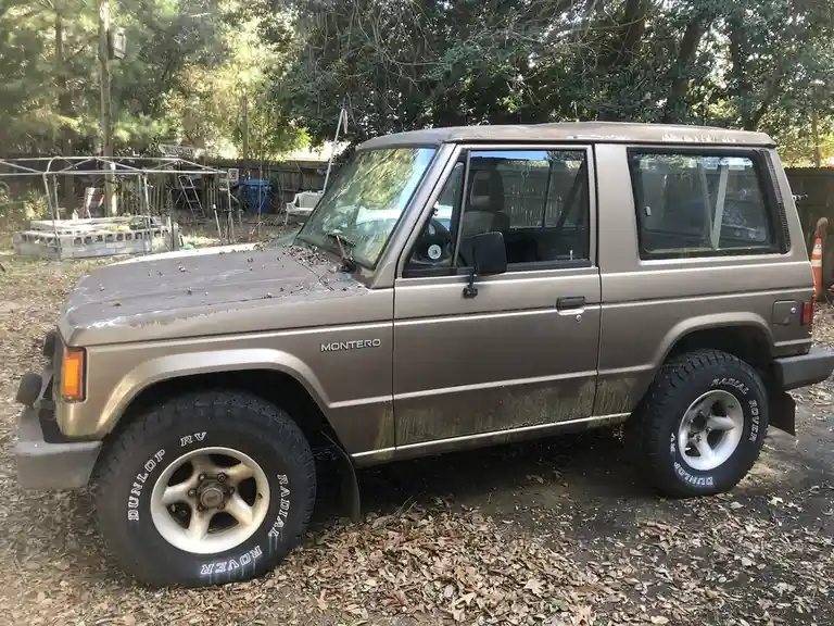 A dusty, brown 1980s-era Mitsubishi Montero SUV is parked on a leaf-covered ground in a backyard, with trees and a fence in…
