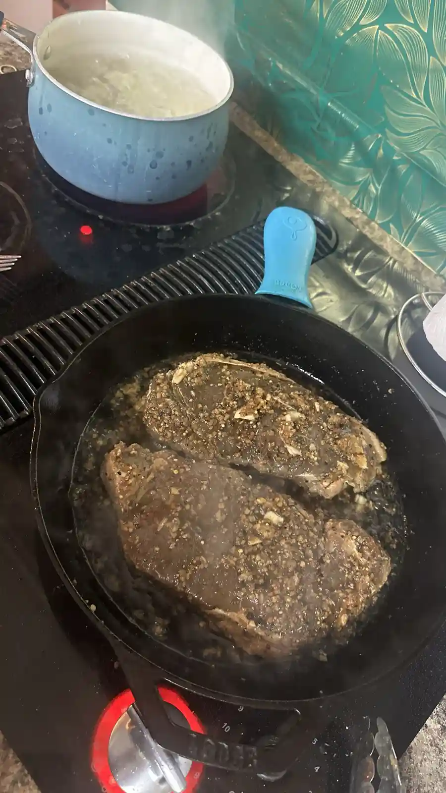 A close-up photo shows two large, seasoned steaks sizzling in a black cast-iron skillet on a stovetop burner, with steam…
