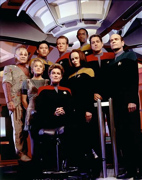 A group photo of the Star Trek: The Next Generation cast in their uniforms, posed in the ship's bridge. The overlaid text…