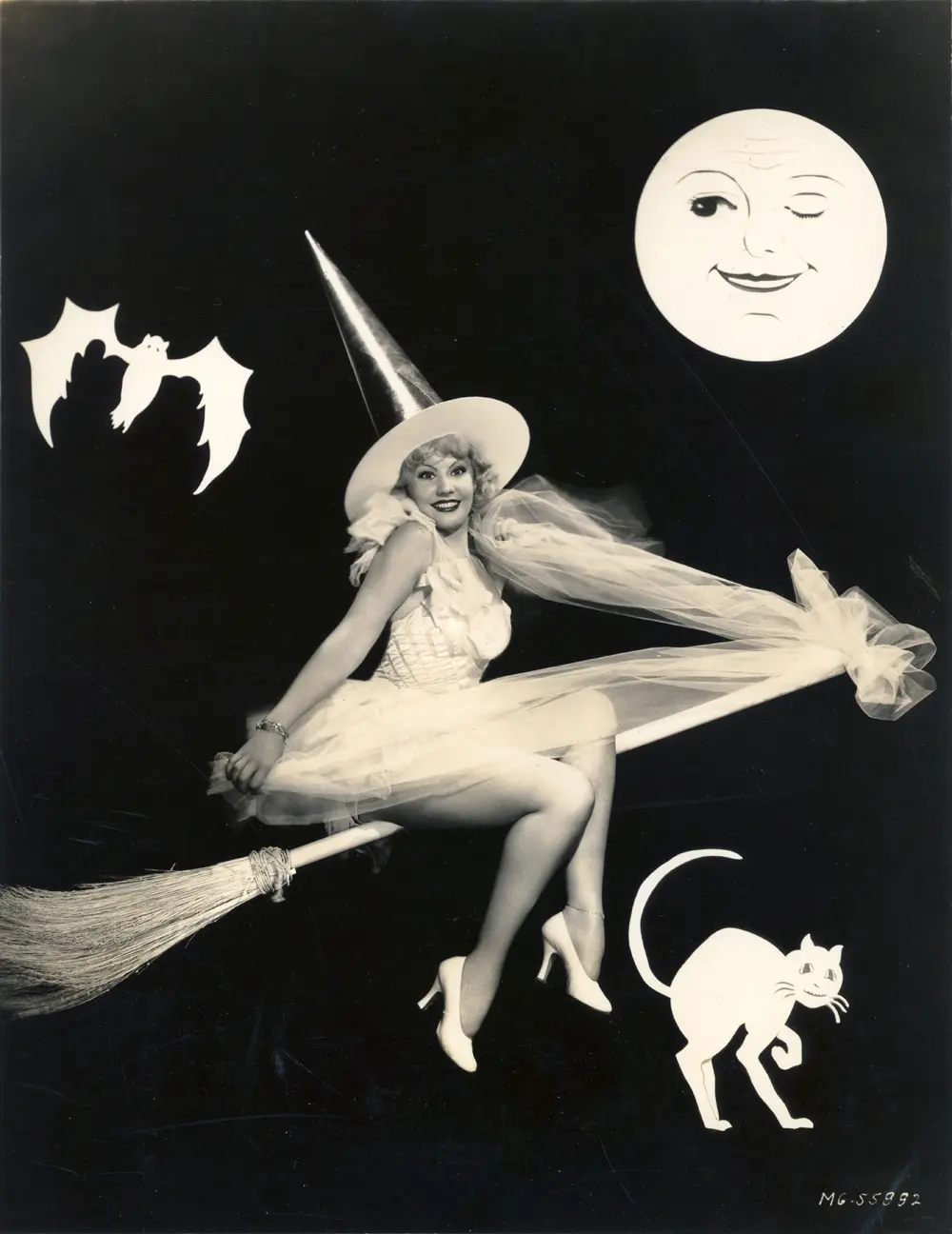 A vintage black-and-white photo shows a smiling woman in a witch costume with a pointed hat and sheer dress, seated on a…