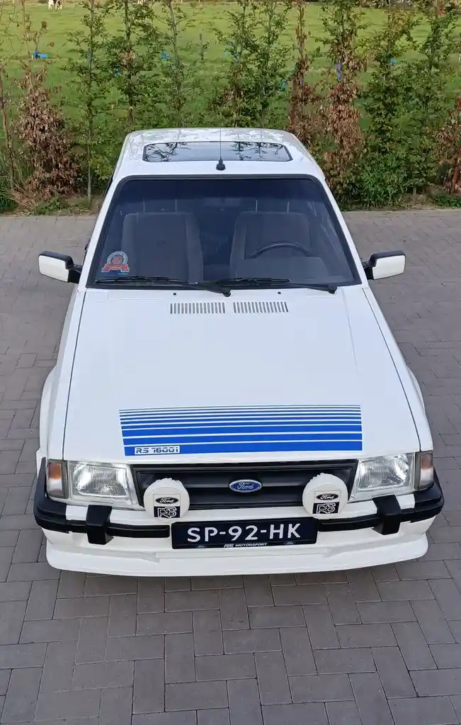 A front-facing photo of a white Ford RS 1600i classic car parked on a paved surface, featuring blue racing stripes on the…