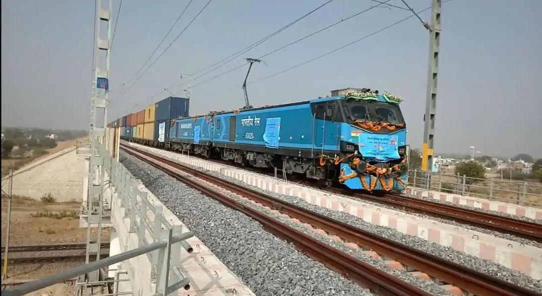 A blue Indian Railways freight train, adorned with orange garlands and banners, travels on a railway bridge under overhead…