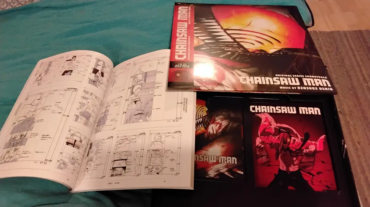 An open book of black-and-white anime storyboards for 'Chainsaw Man' lies next to a vinyl soundtrack box set and two…
