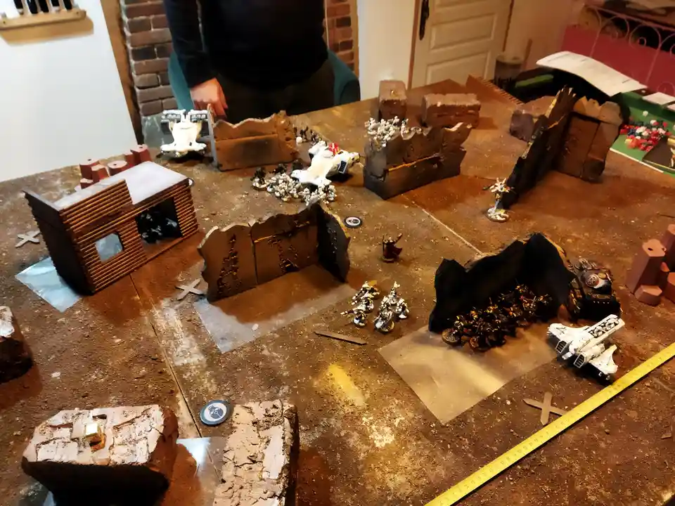 A tabletop wargame setup with miniature figures, terrain pieces like rubble and a small wooden building, and a yellow…