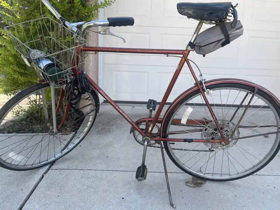 A rust-colored Schwinn Suburban bicycle is parked on a concrete driveway in front of a white garage door, with a wire basket…