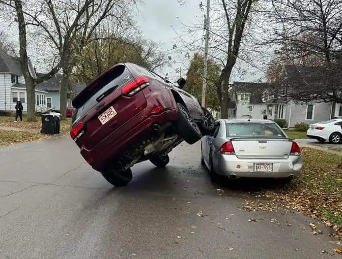 A maroon SUV is visibly tilted at a steep angle, its rear end lifted off the ground and resting against the rear bumper of a…