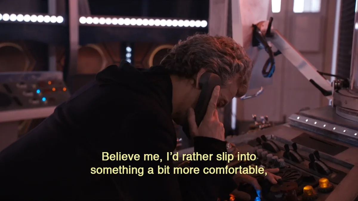 A man with curly gray hair, wearing a black hoodie, is leaning over a control panel in a sci-fi setting while talking on a…